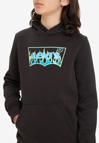 Svart hoodie med en framficka i kängurustil, prydd med en blå och grön elddesign runt "Levi's"-logotypen på bröstet.