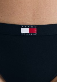 Melnas bikini apakšda ar gludu tekstūru un redzamu Tommy Hilfiger etiķeti, kuras dizains veidots sarkano, balto un zilo krāsu blokos.