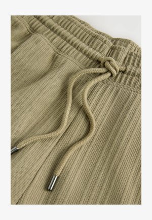 Pantaloni jogger color khaki realizzati in tessuto testurizzato, con vita elasticizzata, coulisse regolabile e dettagli in metallo argentato.