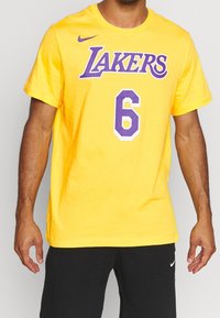 Żółta bawełniana koszulka z krótkim rękawem z fioletowym napisem "LAKERS" oraz numerem "6" w stylizowanym wzorze. Gładka faktura, okrągły dekolt.