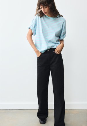 Femme portant une chemise rayée bleu clair et un pantalon noir, debout les mains dans les poches contre un mur blanc uni.