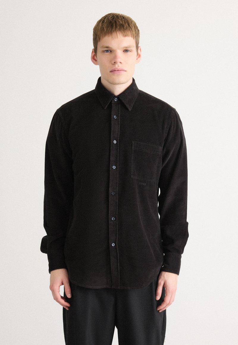 Black corduroy shirt with long sleeves, front pocket, and button-down closure. Features a textured surface and dark blue buttons.