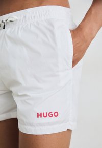Fehér fürdőnadrág rugalmas derékpánttal és zsinórral; oldalzsebével és piros "HUGO" logóval a bal alsó lábszáron.