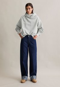 Grijze oversized hoodie met een kap en geborduurde details, gecombineerd met donkerblauwe wijde jeans met opgerolde hems, en beige sneakers.
