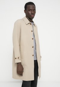 Μπεζ καπαρντίνα από ελαφρύ ύφασμα, με λαιμόκοψη με γιακά, καφέ κουμπιά και πλαϊνές τσέπες. Φορεμένη πάνω από γκρι μπλούζα και σκουρόχρωμο παντελόνι.