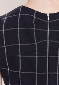 Svart tyg med vitt rutmönster, med en slank halsringning och en centrerad silverdragkedja bak, som ger en elegant design.