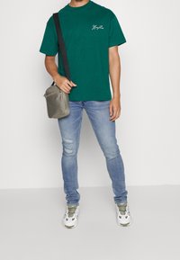 Zelené bavlněné tričko s krátkým rukávem, doplněné světle modrými roztrhanými džíny a šedou křížovou taškou. Bílé a zelené tenisky dotvářejí celkový outfit.