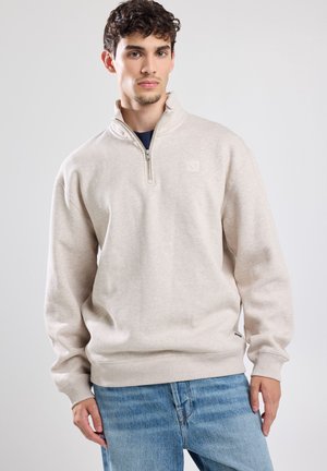 Sudadera de cremallera en color beige claro con cuello alzado, confeccionada en una tela suave. Cuenta con un pequeño logo en el pecho y puños de canalé.