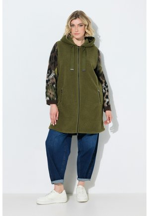 Kvinde stående iført olivengrøn vest med lynlås og hætte over camouflagemønstret langærmet trøje, blå opsmøgede jeans og hvide sneakers.