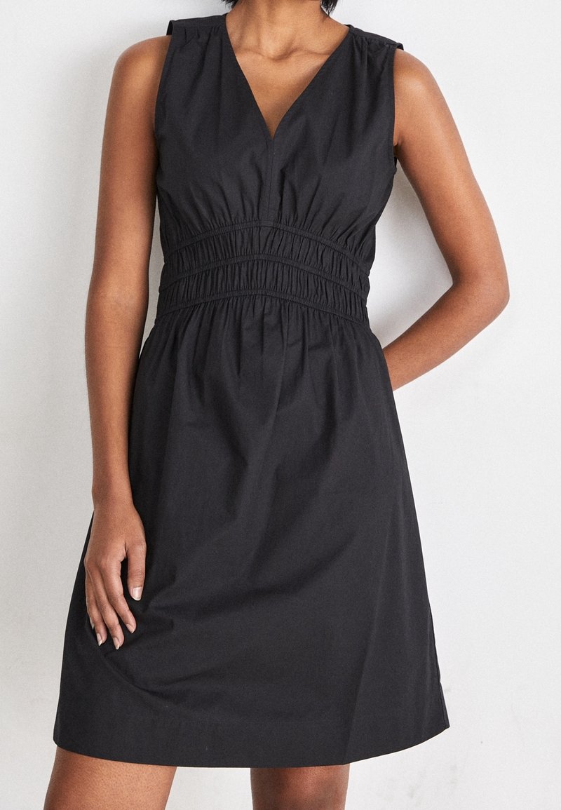 Svart klänning med V-ringning, ärmlös design, draperad midja och svängd kjol. Tillverkad av ett mjukt tyg i en solid färg.