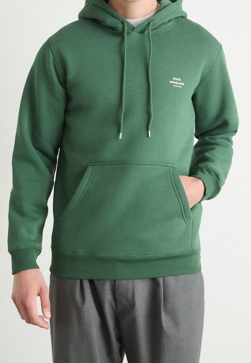Homme portant un sweat à capuche vert avec poche avant et petit logo blanc "Mads Nørgaard Copenhagen" sur la poitrine gauche, associé à un pantalon gris.