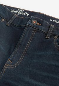 Donkerblauwe denim jeans met contrasterende oranje stiksels, een metalen knoopsluiting en een label met de tekst "NEXT DENIM GOODS CO" op de tailleband.