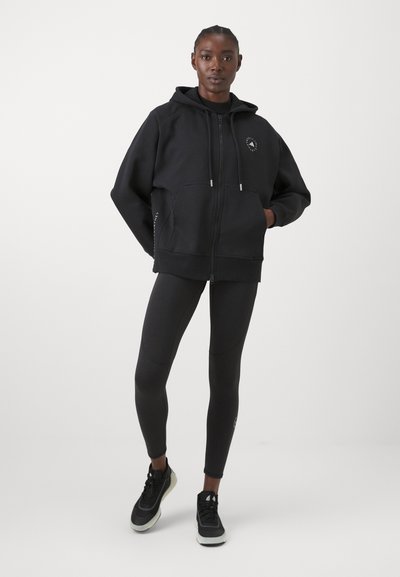 Zwarte rits hoodie met een gevoerde capuchon en voorzakken, gecombineerd met zwarte leggings en zwarte sportschoenen met witte zolen.