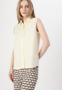 Camisa amarilla sin mangas con botones y color, hecha de una tela suave; combinada con pantalones negros y beige con patrón que presentan diseños ovalados.