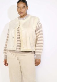 Crème neplederen vest met ronde halslijn, knoopsluiting en zachte textuur, gelaagd over een gestreepte beige en witte trui, gecombineerd met beige corduroy-onderdelen.