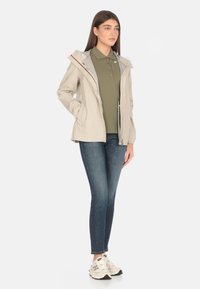 Giacca impermeabile beige con cappuccio, chiusura con zip e polsini elasticizzati, indossata sopra una polo verde, abbinata a jeans blu e sneakers bianche.