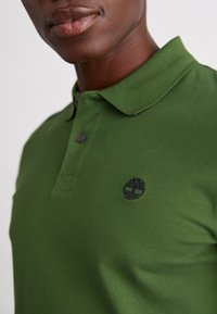 Camisa polo verde feita de tecido texturizado, com colarinho, placket de dois botões e um logótipo bordado em preto no peito.