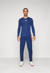 Maglietta sportiva a maniche lunghe blu navy con logo bianco, abbinata a pantaloni blu navy con strisce laterali arancioni e scarpe sportive bianche.