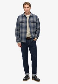 Uomo che indossa una camicia a quadri blu con bottoni sopra un maglione beige, jeans scuri arrotolati, cintura marrone, orologio da polso e scarpe in pelle nere mentre sta in piedi.