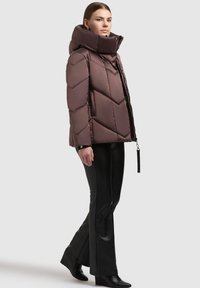 Pruun paks jakk kõrge kraega ja kalasabamustriga, millel on lukk ja silt, kombineeritud mustade liibuvate pükste ja kontsakingadega.