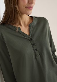 Femme portant un chemisier vert foncé à manches longues avec un col rond et trois boutons noirs sur le plastron avant.