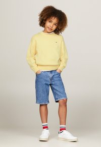 Felpa gialla, pantaloni corti di jeans e sneakers bianche con calzini a strisce rosse e nere. I pantaloni corti sono arrotolati all'orlo per un look casual.