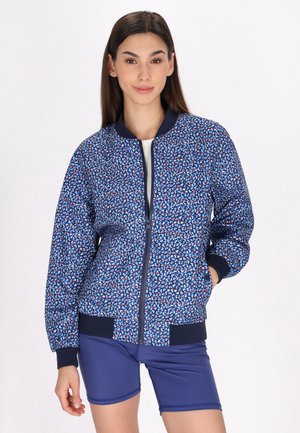 Giacca bomber blu con motivo geometrico multicolore, chiusura con zip, polsini a coste e vita a coste. Indossata con pantaloni corti blu navy.