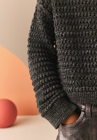 Pull en crochet gris foncé avec un motif texturé, des manches côtelées et un col rond, associé à un pantalon argenté brillant. Objet rond rose en arrière-plan.