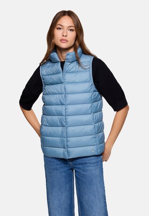 Chaleco acolchado azul claro con cuello alto, costuras horizontales y un emblema de marca en la parte inferior. Llevado sobre una camiseta negra.