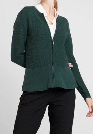 Personne portant un cardigan zippé texturé vert foncé sur une chemise blanche à col et un pantalon noir, debout avec une main légèrement levée.