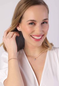 Des boucles d'oreilles couleur argent et un collier délicat avec un pendentif, présentés aux côtés d'un bracelet en or rose, sur un fond neutre.