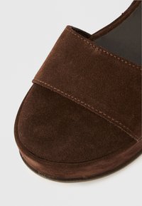 Καφέ σουέντ σανδάλι με φαρδιά λουρίδα, στρογγυλεμένη μύτη και επίπεδη σόλα. Διαθέτει ορατή ραφή κατά μήκος της άκρης. Λείο υλικό.