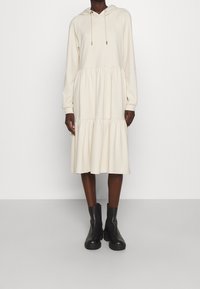 Robe beige à capuche avec un design à étages, des manches longues et des accents à cordon, associée à des bottines noires.