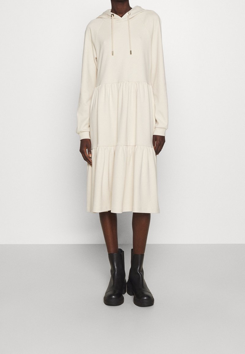 Robe beige à capuche avec un design à étages, des manches longues et des accents à cordon, associée à des bottines noires.