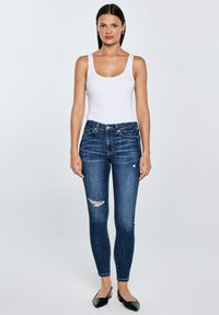 Moteris dėvi baltą marškinėlį be rankovių, tamsiai mėlynas suplėšytas siauras džinsus ir juodus aštriais galiukais plokščius batus, stovinti priešais paprastą šviesų foną.