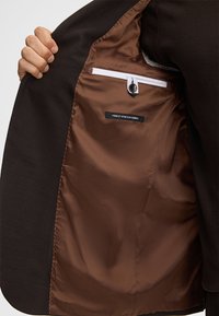 De binnenkant van de bruine jas heeft een gladde bruine voering, een witte ritszak, een zwarte knoop en een label met de tekst "FINEST STRETCH FABRIC."