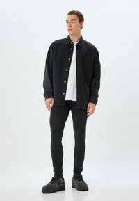Chaqueta de denim negra con bolsillo, cierre de botones y cuello clásico sobre una camiseta blanca. Jeans negros ajustados y zapatillas negras con estampado.
