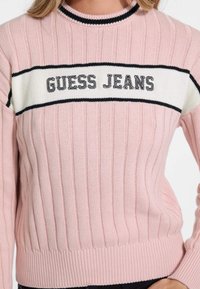 Ljusrosa stickad tröja med ribbat mönster, svart och vit randig band över bröstet med "GUESS JEANS" i svart text.