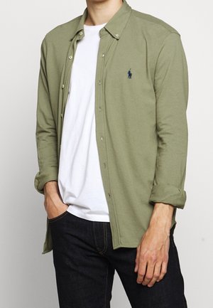 Man met een open olijfgroene lange mouw overhemd over een wit T-shirt en donkere spijkerbroek, linkerhand in de zak, rechterarm ontspannen.