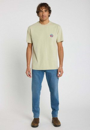 T-shirt beige clair à manches courtes avec un petit graphique coloré sur la poitrine gauche, associé à un jean en denim bleu et des bottes marron.