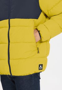 Gul och marinblå pufferjacka med dragkedjestängning, sidofickor med dragkedjor och en logotyp på nederkanten. Mjuk, quiltad textur.