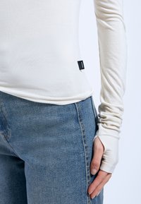 Long-sleeve cream T-shirt with thumbholes, made of soft fabric; paired with high-waisted blue denim jeans featuring stitching details.
