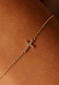 Gouden kettingarmband met een klein kruisje versierd met heldere stenen, gelegen op gebruinde huid. Soepele textuur met delicate schakels.