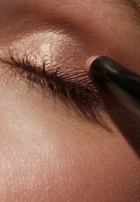 Zbliżenie na oko z nałożonym brązowym cieniowaniem, podczas gdy czarny eyeliner jest stosowany wzdłuż linii rzęs. Widoczna tekstura skóry.