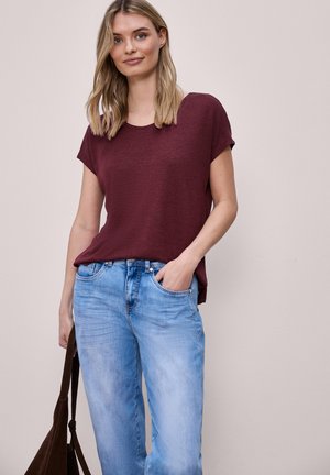 Donna con capelli biondi che indossa una maglietta bordeaux a maniche corte e jeans azzurri, che tiene una borsa marrone scuro in una mano, posa casual.