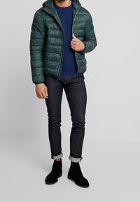 Green puffer jacket with a hood, featuring quilted sections, front zipper, and grey accents. Worn over a blue sweater and dark jeans.