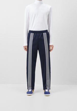Persoon die navyblauwe trainingsbroek met witte strepen draagt, een witte coltrui met lange mouwen en blauwe sneakers met gele strepen, staand tegen een witte achtergrond.