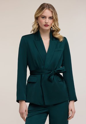 Lyshåret kvinde iført mørkegrøn blazer med bælte og matchende bukser, stylet med guldbelagte øreringe, stående mod en beige baggrund.