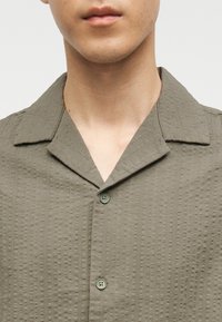 Άνδρας που φοράει υφασμένο πουκάμισο ελιάς με κουμπιά και γιακά τύπου κατασκήνωσης, με ορατό λαιμό και κάτω μέρος προσώπου χωρίς εμφανή έκφραση.