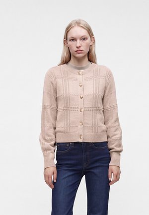 Jeune femme portant un cardigan rose clair texturé boutonné et un jean bleu foncé, debout devant un fond clair uni.